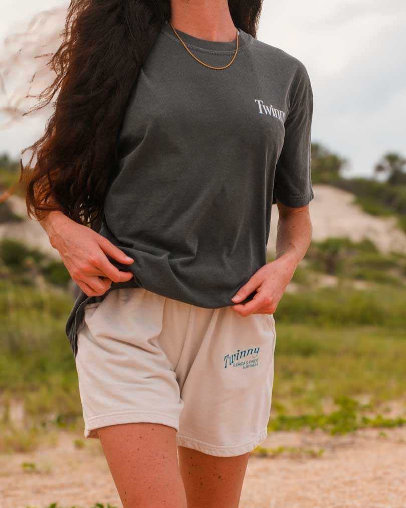 Person wearing a dark gray t-shirt and beige shorts with visible branding outdoors.